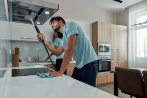 Young caucasian man in uniform cleaning kitchen range hood with steam cleaner while his smiling female colleague washing something in the sink on the background.