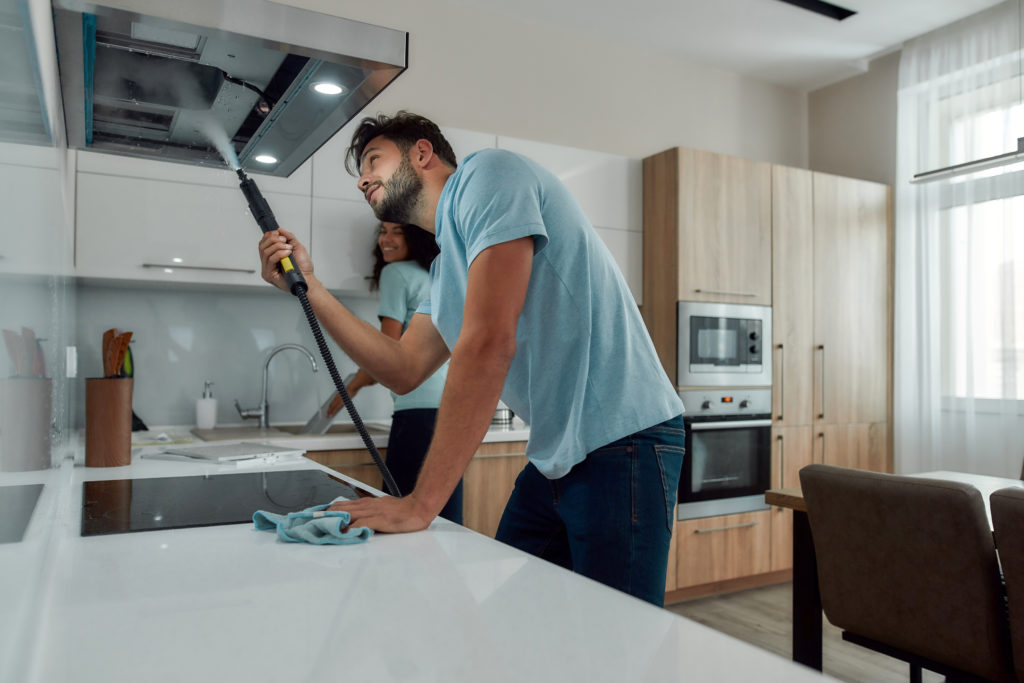 Young caucasian man in uniform cleaning kitchen range hood with steam cleaner while his smiling female colleague washing something in the sink on the background. Cleaning services concept
